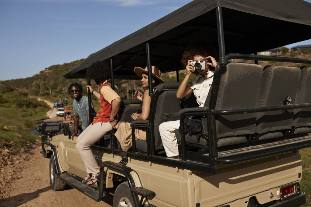 Young woman photographing through camera while sitting with friends in off-road safari vehicle at national wildlife reserve against sky on sunny day